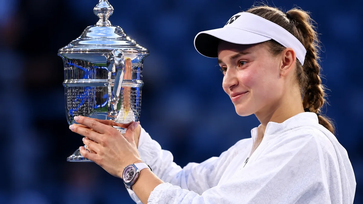 Elena Rybakina smiles as she holds the US Open trophy, wearing a white visor and jacket